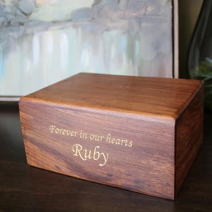 May include: A wooden box with the inscription "Forever in our hearts Ruby" engraved on the front. The box is made of dark wood with a natural grain pattern.