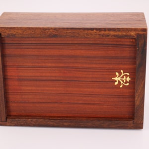 May include: A rectangular wooden box with a hinged lid. The box is made of dark brown wood with a reddish-brown front panel. A small gold-colored decorative emblem is on the front. The box is likely used for storage or display.
