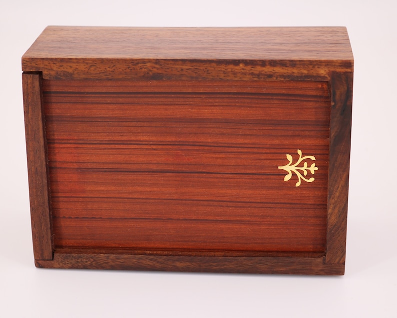 May include: A rectangular wooden box with a hinged lid. The box is made of dark brown wood with a reddish-brown front panel. A small gold-colored decorative emblem is on the front. The box is likely used for storage or display.