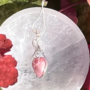 May include: A silver pendant necklace with a teardrop-shaped pink gemstone. The pendant is wire-wrapped and hangs from a delicate silver chain. The necklace rests on a white, round surface surrounded by red and green foliage.