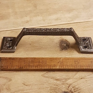 May include: A black cast iron door handle with an ornate design. The handle is approximately 5 inches long and has two holes for mounting.