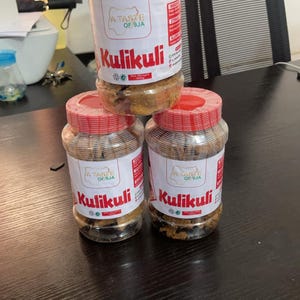 May include: Three clear plastic jars of Kulikuli, a Nigerian snack, with red lids and white labels. The labels feature the text "A Taste of 9ja" and the product name. The jars are stacked on a dark wooden surface.