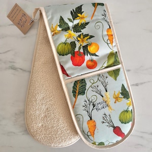 May include: Double oven glove with a light blue vegetable print featuring tomatoes, carrots, and squash. The backing is beige terry cloth. A tag says "Emily Packer".