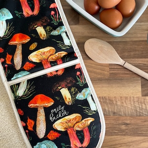 May include: A pair of black oven mitts with a white trim and a mushroom pattern. The mitts have the text "emily hacker" written on them. The mitts are on a wooden surface with a bowl of eggs and a wooden spoon.