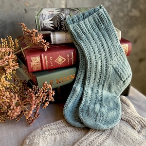 Puede incluir: Dos pares de calcetines de punto, uno en azul claro y otro en beige claro. Los calcetines azules son acanalados, mientras que los calcetines beige tienen un diseño estampado. Los calcetines se exhiben con una pila de libros antiguos y flores secas.