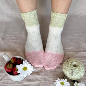 May include: Hand-knitted socks with a white body, light green cuffs, and pink toes. The socks are shown with a cup of strawberries, daisies, and a ball of yarn.