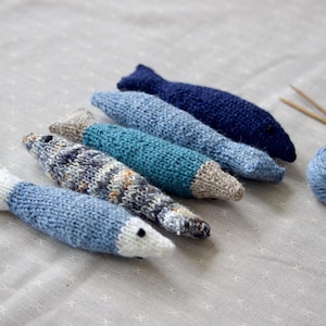 May include: A collection of six knitted fish toys in various shades of blue, with white and gray accents. A ball of blue yarn and knitting needles are also present. The fish are arranged on a light gray surface.