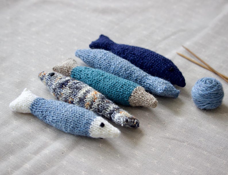 May include: A collection of six knitted fish toys in various shades of blue, with white and gray accents. A ball of blue yarn and knitting needles are also present. The fish are arranged on a light gray surface.