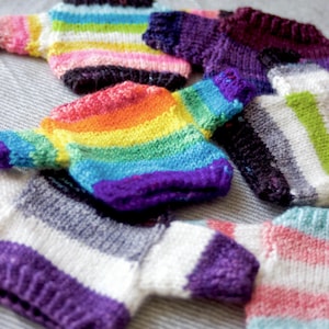Op de afbeelding: Een verzameling van vijf gebreide truien in verschillende kleuren en patronen, waaronder regenboog, roze en witte strepen, grijs en witte strepen en paars en witte strepen.