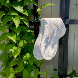 Op de afbeelding: Een paar witte sokken met roze stippen die aan een zwart metalen hek hangen. De sokken zijn gebreid en hebben een zachte, textuur.