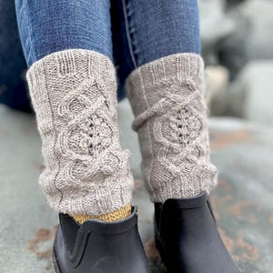 May include: A pair of light gray knitted leg warmers with a cable knit design and a leaf pattern. The leg warmers are worn over yellow socks and black rubber boots. The person is wearing blue jeans.