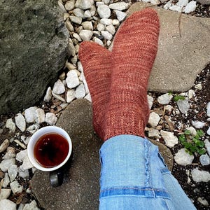 May include: A pair of rust-colored knitted socks with a subtle texture, worn with blue jeans. A cup of tea sits on a stone surface.