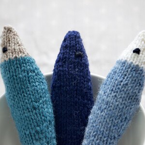 May include: Three handmade knitted fish toys in shades of blue, with white and tan accents. Each fish has a small black eye. The toys are arranged in a white bowl, showcasing the handcrafted details and textures of the yarn.