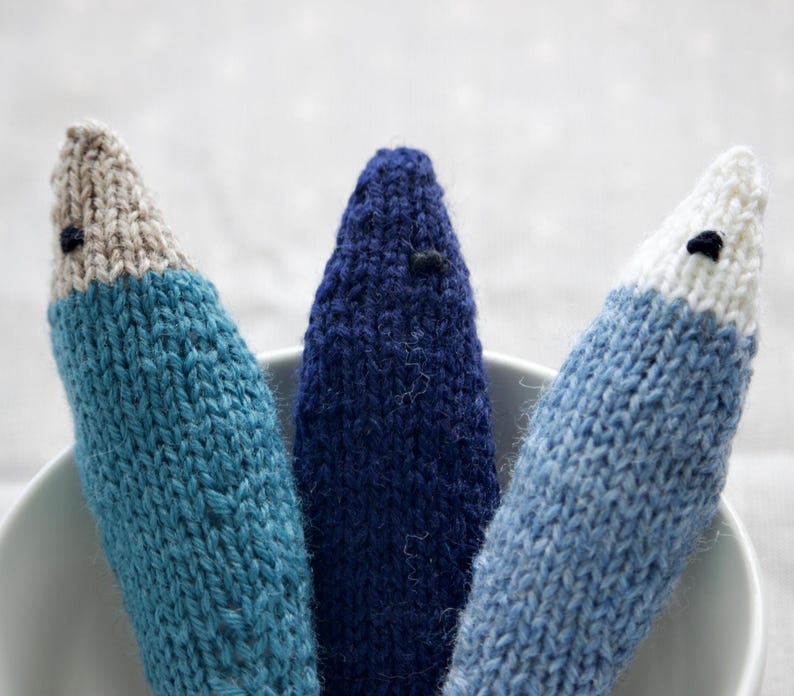 May include: Three handmade knitted fish toys in shades of blue, with white and tan accents. Each fish has a small black eye. The toys are arranged in a white bowl, showcasing the handcrafted details and textures of the yarn.