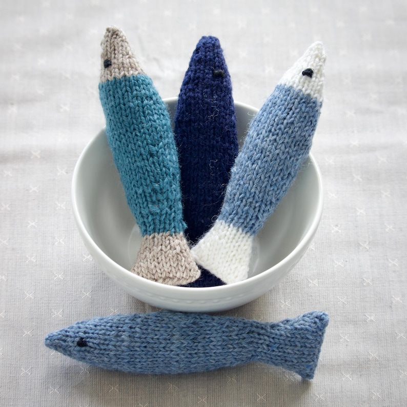 May include: A collection of knitted fish toys in shades of blue, teal, and white, with a beige or white accent at the base. One fish lies outside a white bowl containing three upright fish. Each fish has a small black eye.