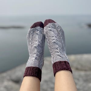 May include: A pair of gray cable-knit socks with dark burgundy cuffs and toes. The socks are patterned with a repeating cable design. The image shows the socks against a blurred background of water and sky.