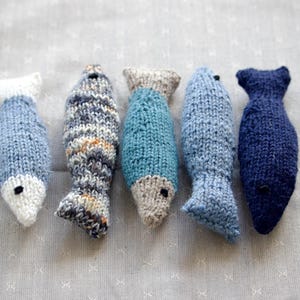 May include: Five hand-knitted fish toys in shades of blue, gray, and white. Each fish has a unique pattern and design, with black bead eyes. The fish vary in size and shape, perfect for a child's toy or home decor.