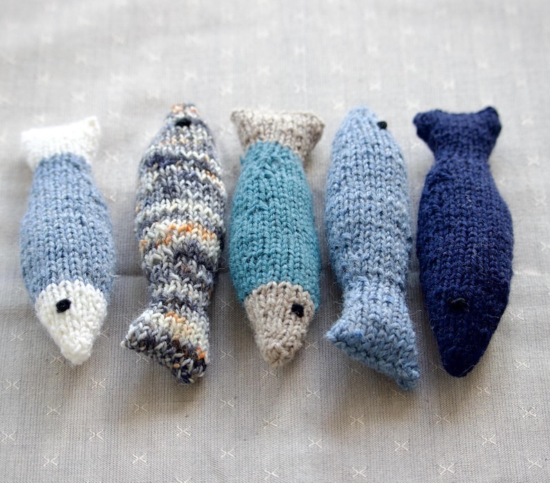 May include: Five hand-knitted fish toys in shades of blue, gray, and white. Each fish has a unique pattern and design, with black bead eyes. The fish vary in size and shape, perfect for a child's toy or home decor.