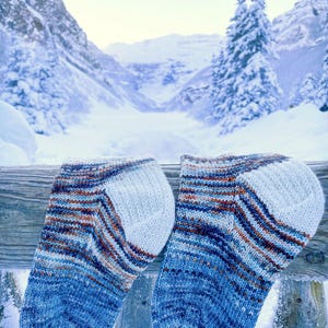 May include: Two blue and brown striped socks with white heels and toes, hanging on a wooden fence against a snowy mountain backdrop.