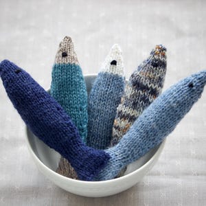 May include: A white bowl filled with six knitted fish toys in various shades of blue, gray, and white. Each fish has a small black eye and a tail fin. The fish are arranged in a fan shape, with their heads pointing upwards.