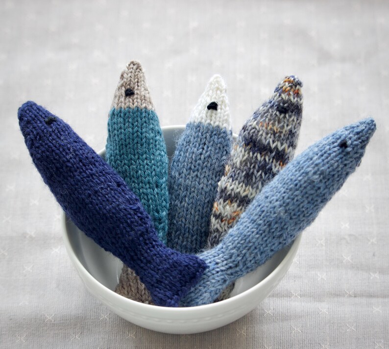 May include: A white bowl filled with six knitted fish toys in various shades of blue, gray, and white. Each fish has a small black eye and a tail fin. The fish are arranged in a fan shape, with their heads pointing upwards.