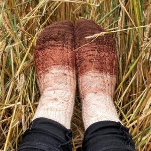 May include: A pair of hand-knitted socks featuring a cable knit pattern. The socks display a gradient color transition, shifting from a dark brown at the toes to a light beige at the cuffs. The knit texture is consistent across both socks.