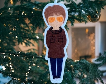 May include: A felt Christmas tree ornament shaped like a person wearing sunglasses, a brown jacket, and dark blue pants. The ornament has a white border and a white string for hanging. The ornament is hanging on a Christmas tree.