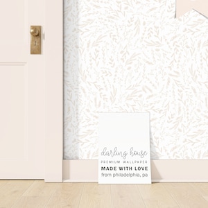 May include: A white wall with a delicate floral pattern in a light beige color. A white sign with the text "darling house PREMIUM WALLPAPER MADE WITH LOVE from philadelphia, pa" is on the floor in front of the wall.