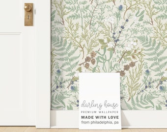 May include: Wallpaper featuring a botanical pattern of green ferns, brown pinecones, white berries, and blue sprigs on a cream background. A white sign in front displays the text: darling house PREMIUM WALLPAPER MADE WITH LOVE from philadelphia, pa.