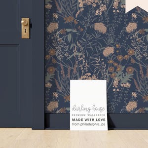 May include: Dark blue floral wallpaper with a botanical design featuring beige, brown, and green flowers and foliage. A white sign in front of the wallpaper reads "darling house PREMIUM WALLPAPER MADE WITH LOVE from philadelphia, pa."