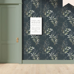 May include: A dark blue wallpaper with a repeating pattern of green leaves and gold accents. The wallpaper is hung in a room with a green door and light wood floors. A sign hanging on the wall reads "Darling House Premium Wallpaper Made with Love from Philadelphia, PA."