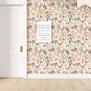May include: Floral wallpaper with a repeating pattern of pink, brown, and yellow flowers on a white background. The text on the sign reads "DARLING HOUSE premium WALLPAPER MADE WITH LOVE from PHILADELPHIA, PA". A white door and wooden floor are also visible.
