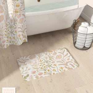May include: A rectangular bath mat with a floral pattern in soft pink, yellow, and green hues. The mat is placed on a light wood floor, near a white bathtub and a matching floral shower curtain. A metal basket holds white towels.