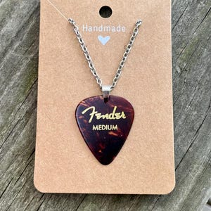 May include: A Fender guitar pick pendant necklace. The pendant is a brown tortoiseshell color with the Fender logo and "MEDIUM" in gold lettering. The necklace has a silver chain and is displayed on a brown card that says "Handmade".
