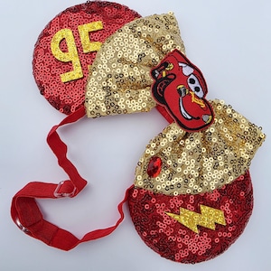 Op de afbeelding: Een paar rode oren met lovertjes, voorzien van een gouden strik met lovertjes, het glinsterende gele getal 95, een gele glinsterende bliksemschicht, een rode edelsteen en een geborduurde patch van een rode raceauto. De oren zijn verbonden door een rode elastische band.