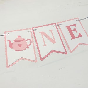 May include: Pink and white banner with the word "ONE" and a pink teapot with a heart on it. The banner is strung with white string.
