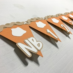 May include: A festive banner featuring orange pie slice-shaped pennants with white accents and brown crust detailing. The banner includes the letters "NB" and numbers 1, 2, 3, and 4, perfect for a birthday celebration.