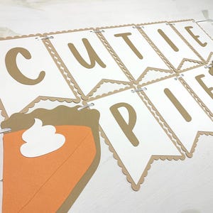 May include: A banner with the words "CUTIE PIE" in brown letters on white pennants. The pennants have a scalloped edge and are strung together with a white ribbon. Two pie slice cutouts are included.