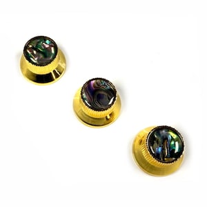 May include: Three gold-colored knobs with a round, iridescent abalone shell inlay. The knobs have a ridged edge and a flat base. The abalone shell inlay features a mix of green, blue, and purple hues.