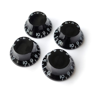 May include: Four black guitar knobs with white numbers and markings. The knobs are designed for use on electric guitars.