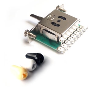 May include: A silver guitar switch with a green circuit board and a black, white and cream knob. The switch has a lever and a series of metal contacts.