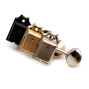 Può includere: Primo piano di tre meccaniche per chitarra nere, dorate e argentate. Ogni meccanica ha una piastra rettangolare con linee verticali e una parte superiore decorativa. La meccanica argentata ha una chiavetta. Lo sfondo è bianco.