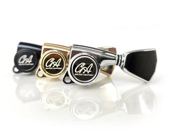 May include: Close-up of four guitar tuning machines in black, gold, and silver. Each machine has a circular face with the letters "CA" and the words "Guitar Anatomy". The tuning machines are on a white background.