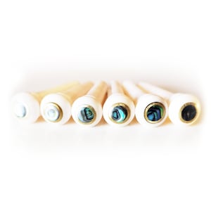 May include: A set of seven guitar bridge pins. Each pin has a white head with a decorative abalone inlay and a brass ring. The pins are made of light-colored wood.