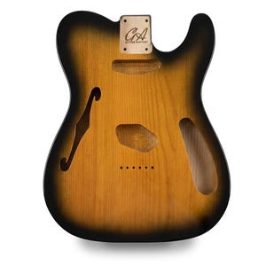 Könnte beinhalten: Ein unfertiger E-Gitarrenkorpus mit Sunburst-Lackierung, der von einem dunklen schwarzen Rand zu einer naturfarbenen Holzmitte übergeht. Er verfügt über ein F-Loch, eine Halstasche mit dem Text GA GUITAR ANATOMY sowie Aussparungen für Tonabnehmer und Brücke.
