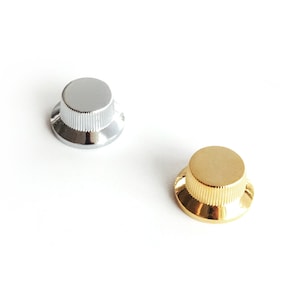 Puede incluir: Dos botones de guitarra, uno plateado y otro dorado. Ambos botones tienen un diseño elevado y acanalado.