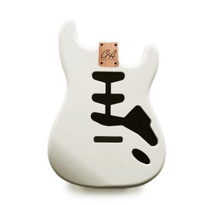 Könnte beinhalten: Ein weißer E-Gitarrenkorpus mit schwarzen Aussparungen für Tonabnehmer und Bedienelemente. Ein kleines Holzstück mit den Buchstaben "GA" und den Worten "GUITAR ANATOMY" ist oben befestigt. Der Gitarrenkorpus hat eine glatte, glänzende Oberfläche.