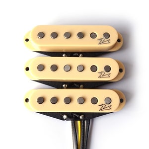 Könnte beinhalten: Drei cremefarbene E-Gitarren-Tonabnehmer mit schwarzen Montage-Ringen und jeweils sechs silbernen Polschuhen. Die Tonabnehmer haben ein schwarz-weißes Logo mit dem Text "Seymour Duncan" darauf.