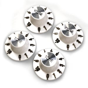 Może przedstawiać: Cztery białe gałki do gitary z czarnymi cyframi i napisami. Dwie gałki są oznaczone jako "VOLUME", a dwie jako "TONE".