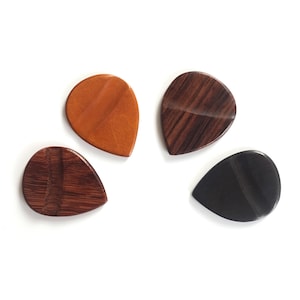Könnte beinhalten: Vier Gitarrenplektren aus Holz. Die Plektronen haben die Form eines Tropfens und verschiedene Holzmaserungen. Ein Plektron ist braun, eines ist schwarz und zwei sind rotbraun.